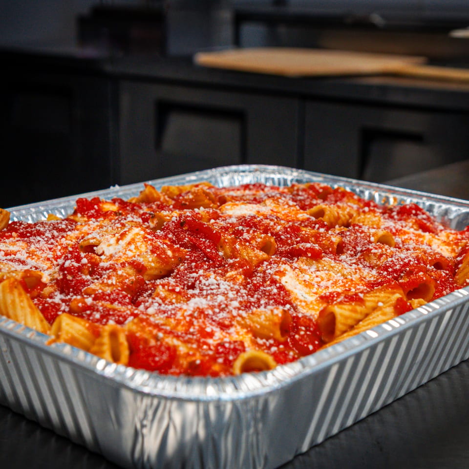 Baked rigatoni pasta covered in tomato sauce and grated cheese, served in a disposable aluminum tray on a restaurant counter.
